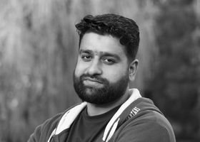 A serious man with a beard in a hoodie poses outdoors. Black and white portrait.